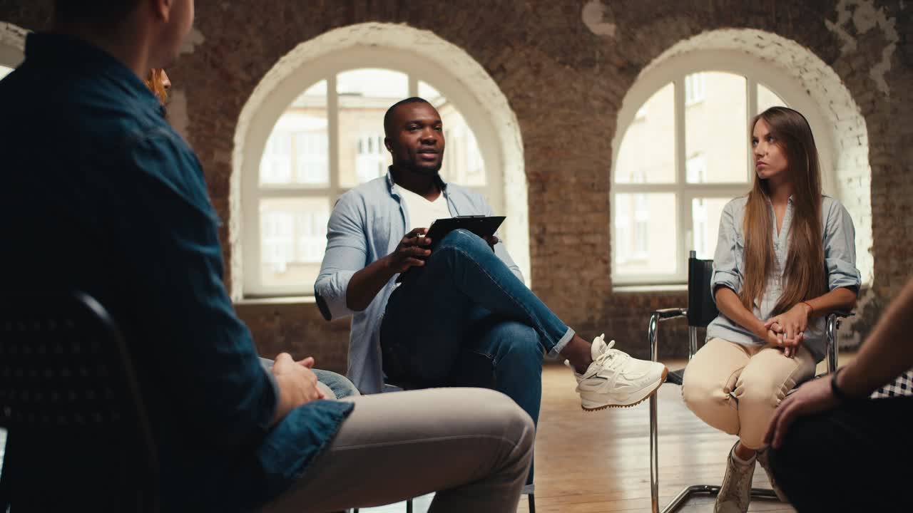 A black man, a psychologist, in jeans, interviews group therapy participants who are sitting in a circle in a brick hall
