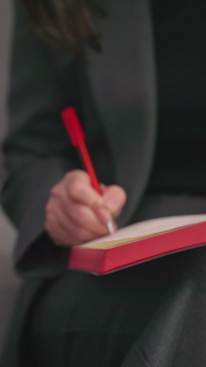 Businesswoman writing in red notebook with pen in hand, sitting in modern office, gray suit, focused on planning, decision-making, productivity, workspace, corporate environment