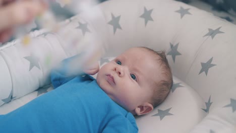 mother plays with cute kid in blue holding rattle over crib