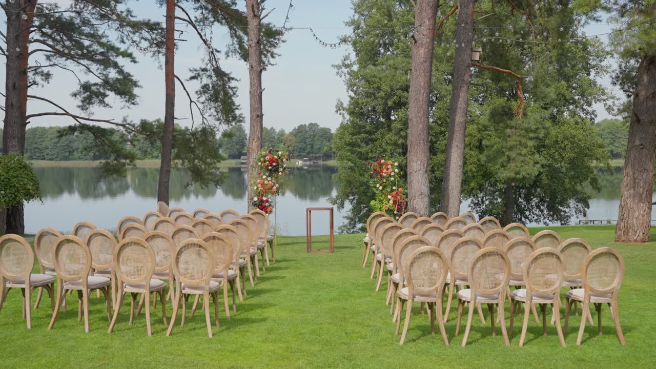 Scena idilliaca di matrimonio all'aperto con sedie vuote elegantemente disposte su un prato verde lussureggiante in riva al lago sotto un cielo sereno. Ambiente tranquillo incorniciato da alberi alti e decorazioni floreali.