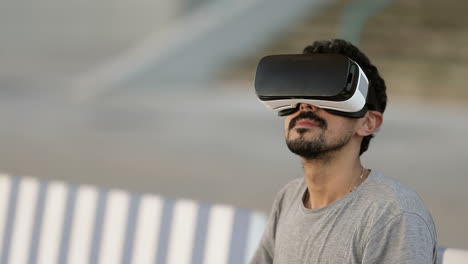 Young-Arabic-man-with-dark-curly-hair-and-beard-in-grey-T-shirt-sitting-in-park-on-striped-bench-in-virtual-reality-glasses