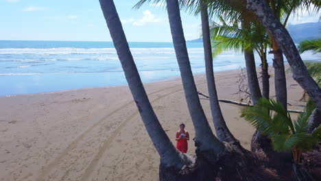 low aerial view of a female tourist in orange outfit flying the drone through some palm trees on tropical beach in costa rica