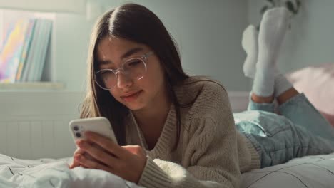 caucasian teenage girl browsing phone while lying on front in bed