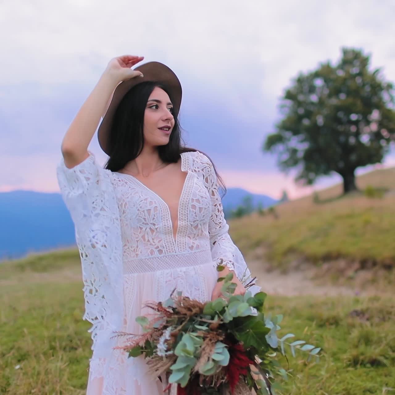Young bride on mountains background. Beautiful lady in white wedding dress holding flowers and is taking off her hat in nature.
