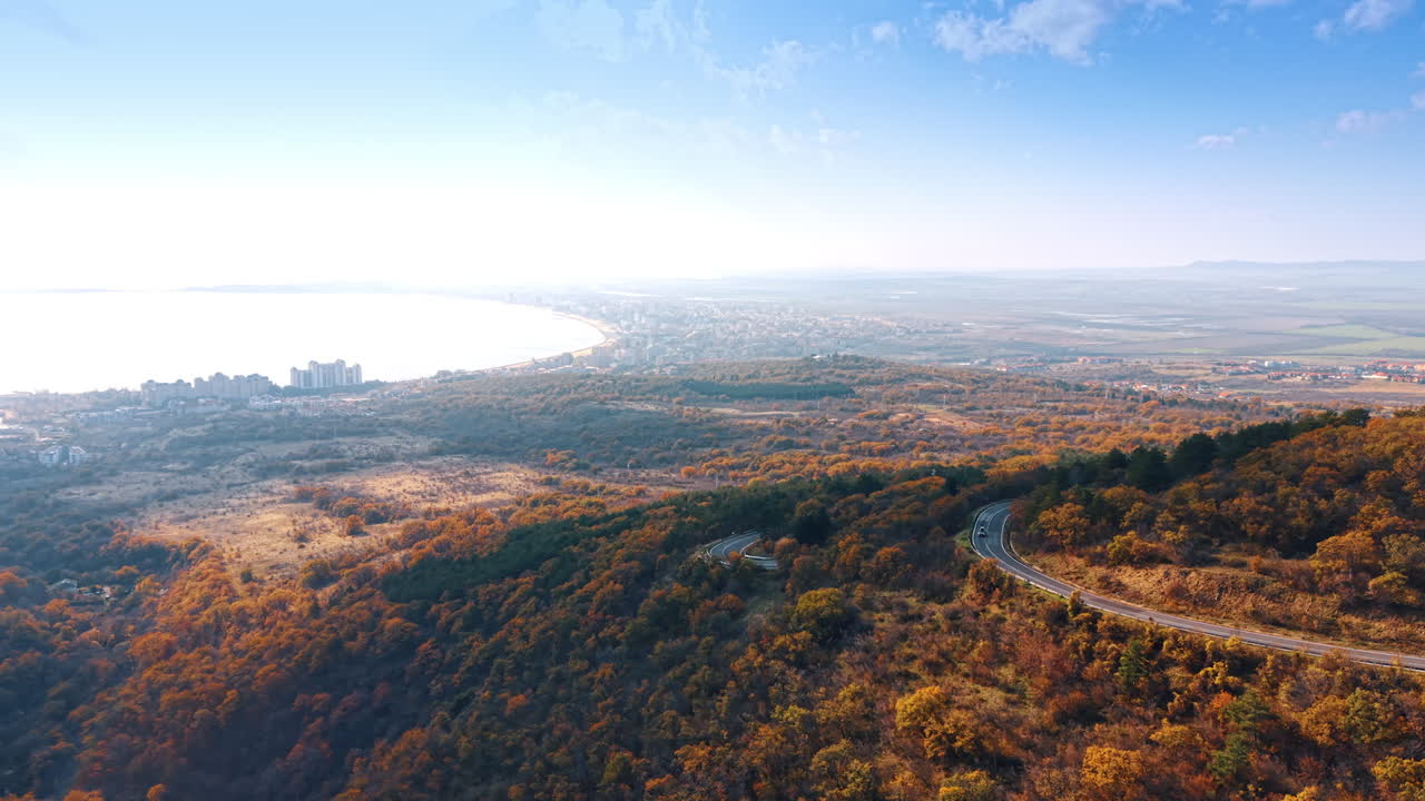 Autumn Mountain Landscape with Coastal City and Road. Stunning autumn panorama showcasing dense forest hills, winding road, coastal city, and lake under clear blue sky