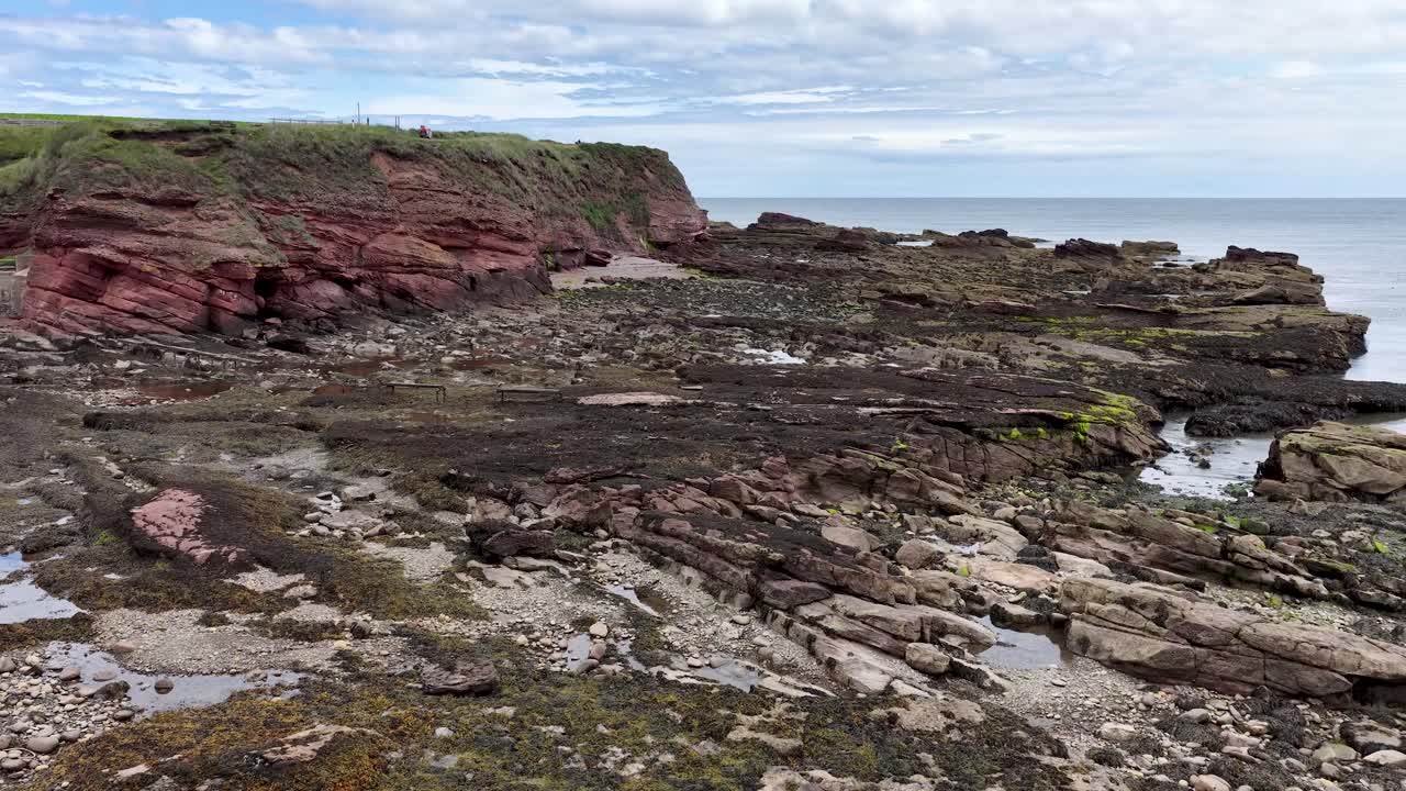 Camera slowly pans across red sandstone cliffs, rocky shore, and tidal pools under soft daylight