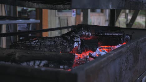 pan around outdoor barbecue fire pit with orange flames flickering from timber charcoal wood with red hot ember bed underneath as ember, smoke and ash fly off