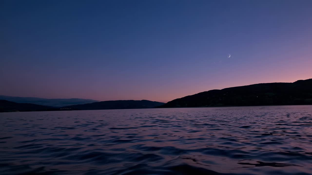 A serene video of a twilight lake scene with a crescent moon, captured from a low-angle perspective