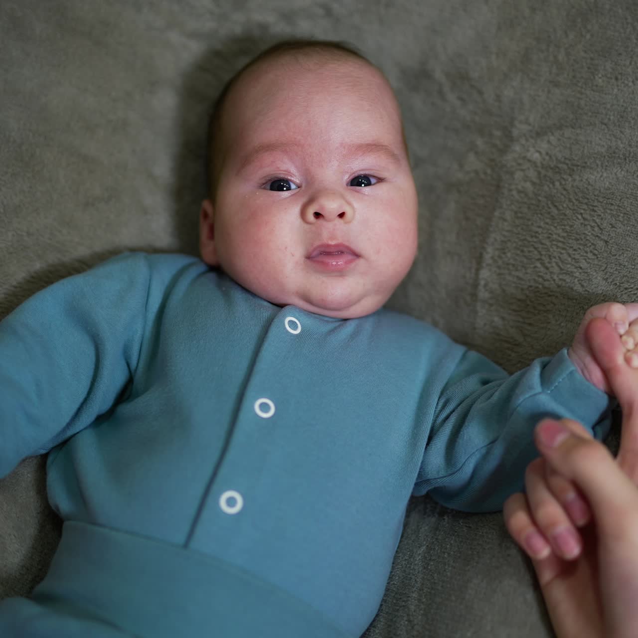Bayi tenang yang mengantuk berbaring di tempat tidur, memegang erat jari ibunya. Bayi laki-laki mungil yang baru lahir mengenakan pakaian biru menguap manis sambil menatap kamera. Tampak atas.