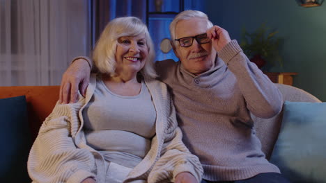 happy old senior elderly family couple hugging, laughing, smiling looking at camera at home sofa