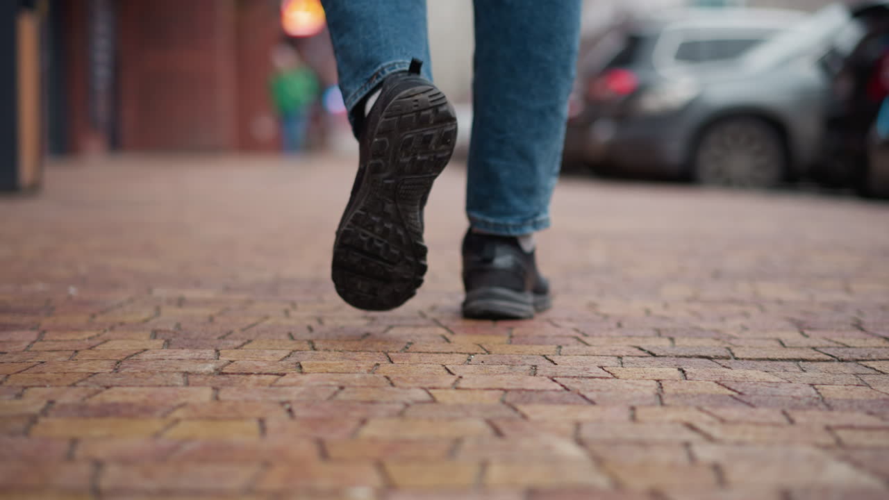 Back leg view of man wearing black winter coat and jeans walking on tiled walkway beside parked cars and urban buildings with focus on boot sole pattern and motion during cold city day scene
