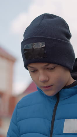 niño con gorra negra y chaqueta azul paseando con la cabeza inclinada pensativamente, fondo borroso con edificios, postes de luz y árboles