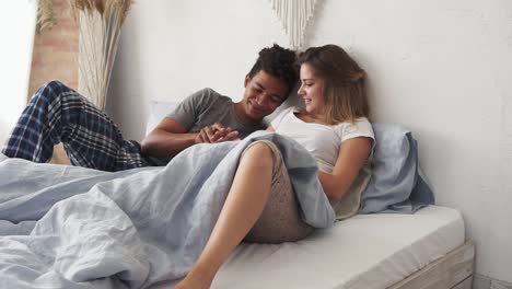 Young cute multi ethnic couple lying in bed and talking. Handsome african man is holding his caucasian girlfriend&#039;s hand and