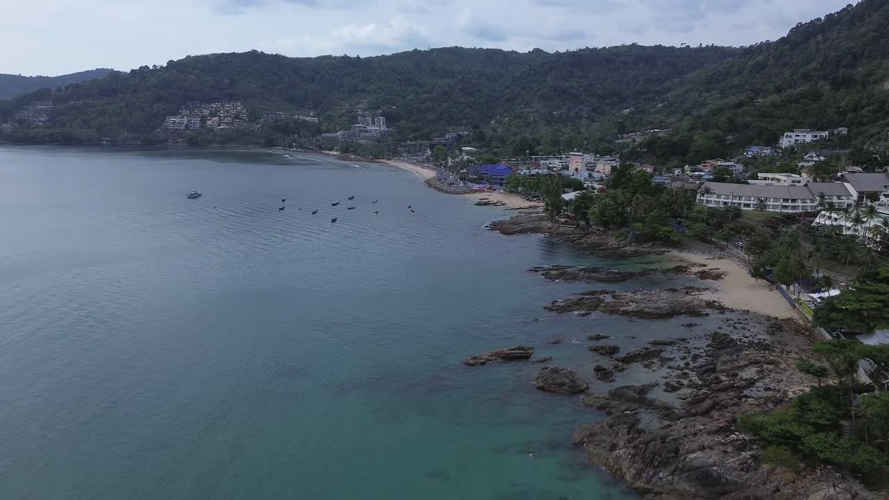 Drone zooms out from coastline revealing buildings and boats on a calm sea under a cloudy sky