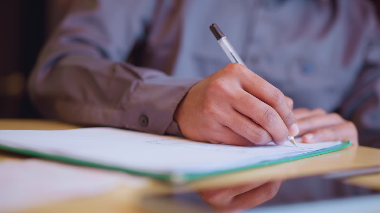 Close-up of male hand in grey shirt drawing detailed sketch on white paper with black pen while seated at wooden desk, focused on creating visual design, surrounded by soft indoor lighting and smartphone