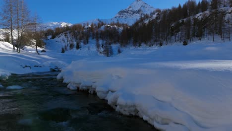 drone flying low over stream flowing through snow covered and frozen valmalenco on sunny day, alps in italy