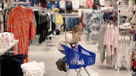 Little-girl-is-having-fun-in-a-clothes-store,-riding-a-shopping-cart