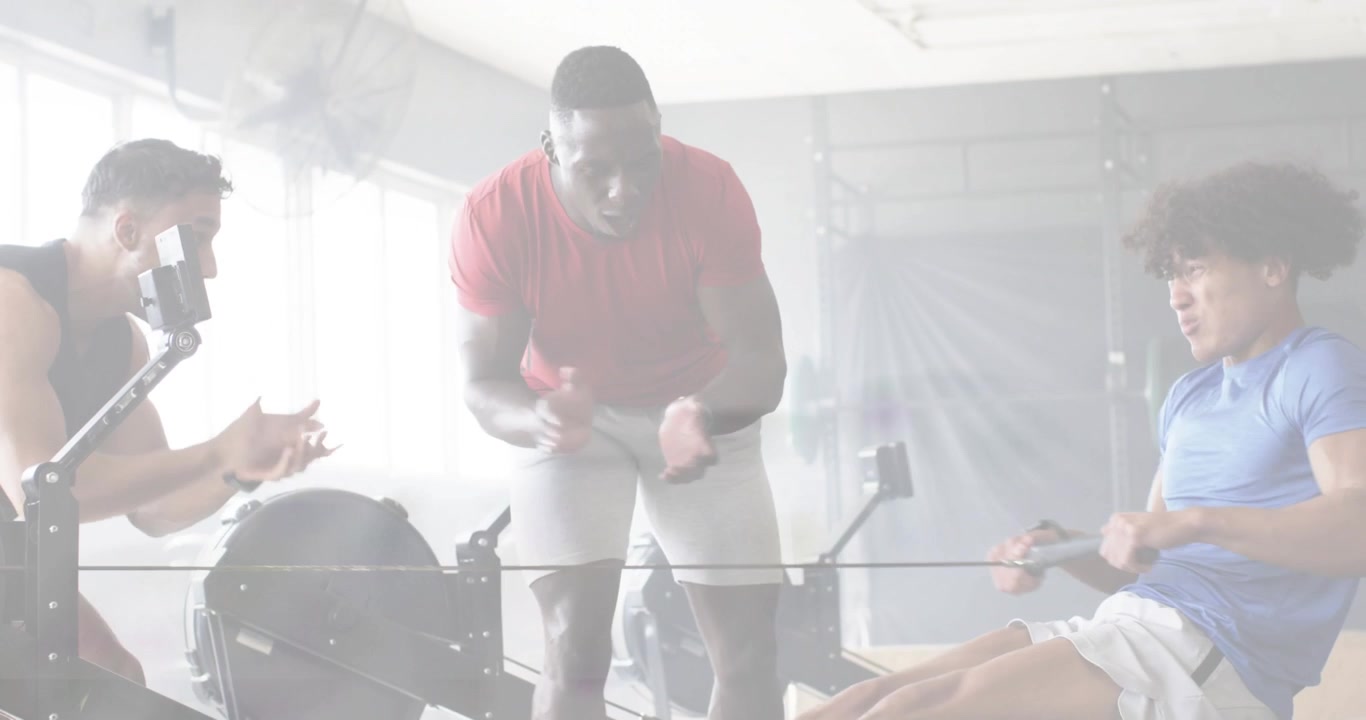 Coach cueing start as male athlete rowing on rower for fitness feedback, teammate holding phone