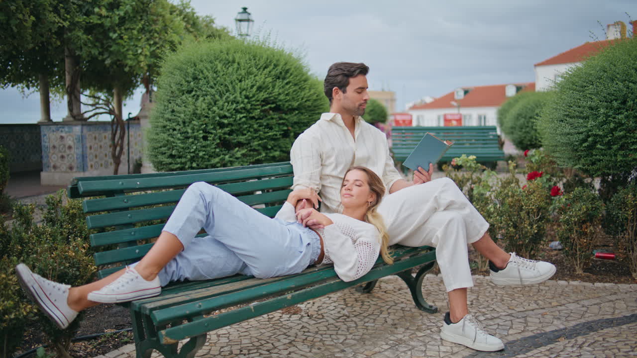 새로 결혼 한 부부 가 공원 에서 함께 휴식 하는 벤치. 갈색 을 입은 남자 가 큰 소리 로 책 을 읽고 있다