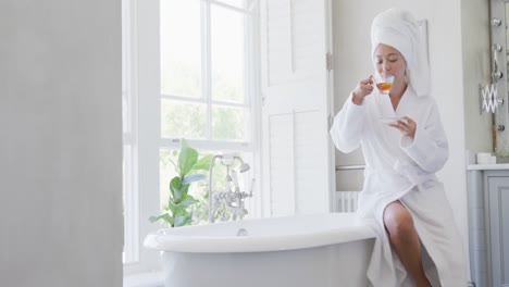 Happy-asian-woman-sitting-on-bathtub-and-drinking-tea-in-bathroom,-in-slow-motion