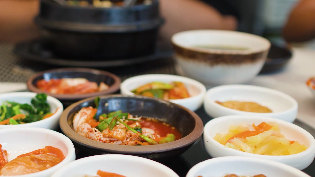 Kamera close-up bergerak melintasi berbagai mangkuk kecil lauk pauk tradisional Korea dan bulgogi.