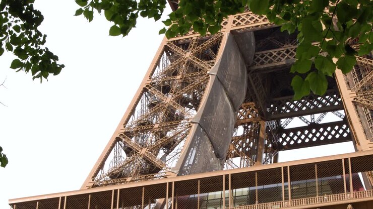 Inquadratura statica dal basso della struttura in ferro battuto della Torre Eiffel, incorniciata da fogliame verde.