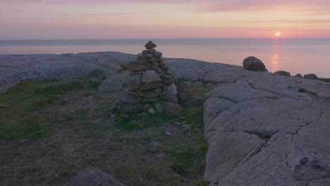 Balancing-rock-sculpture-stands-on-the-top-of-a-mountain,-overlooking-the-calm-ocean-with-a-beautifully-rich-pink-sunset-and-clear-sky