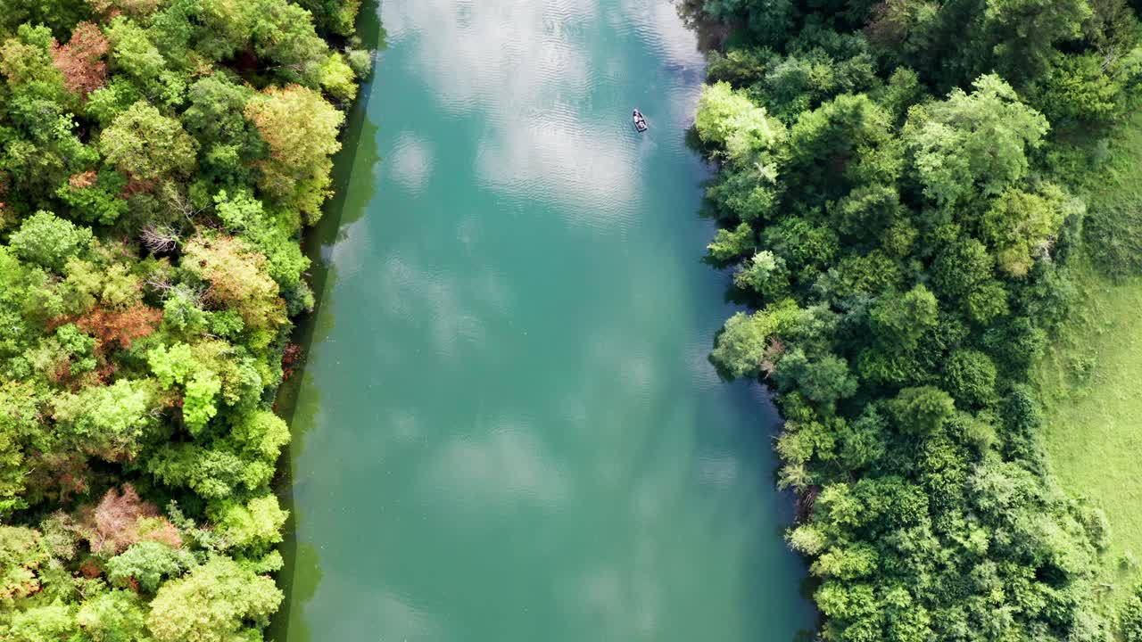 Clouds reflected in deep green-blue water of river through wooded area with different colored trees, aerial drone