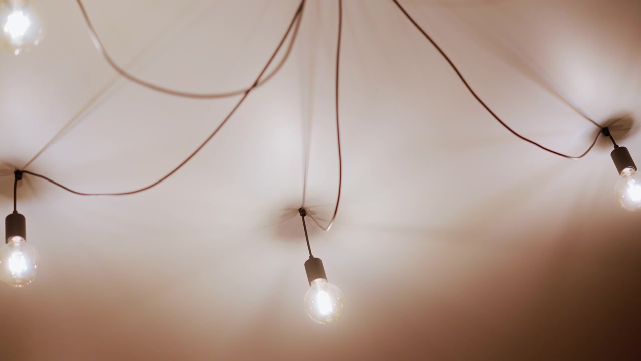 Primo piano di più lampadine a vista appese al soffitto con fili visibili, che brillano delicatamente e creano un'illuminazione ambientale calda. Le lampadine sono distanziate uniformemente su uno sfondo semplice.
