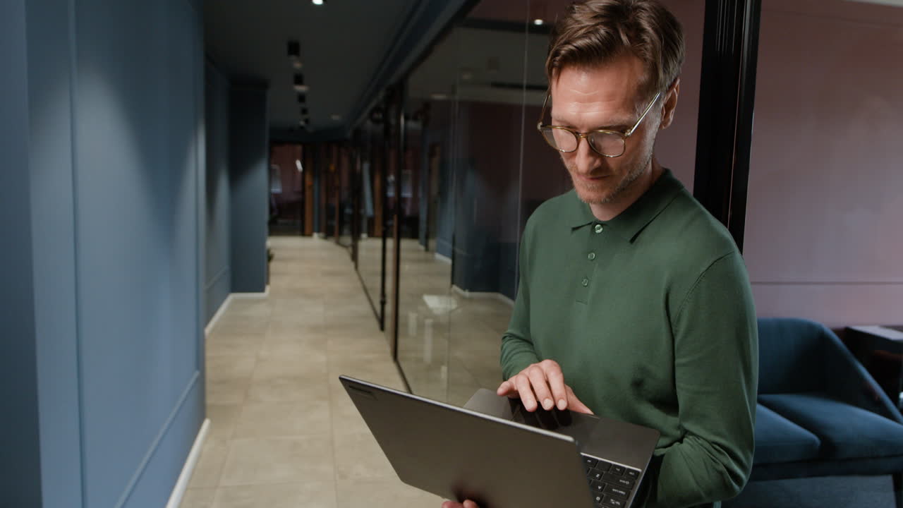 Man standing and working on a laptop in an office hallway
