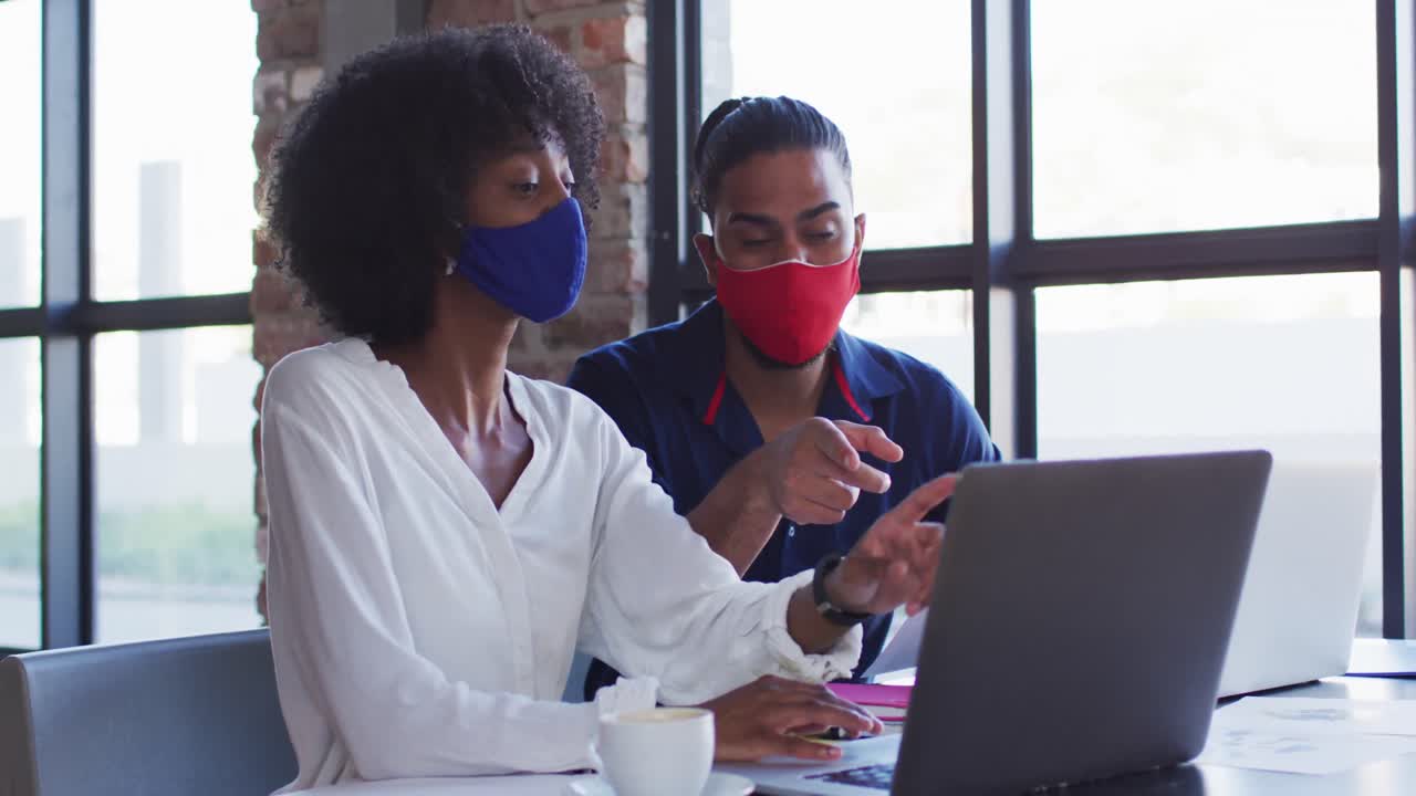 Diverse couple wearing face masks sitting in cafe using laptops and talking