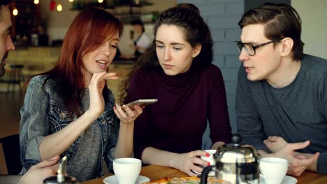 cheerful young men and women close friends are using smartphone and talking while having lunch in nice cafe. dining, modern technology and friendship concept.