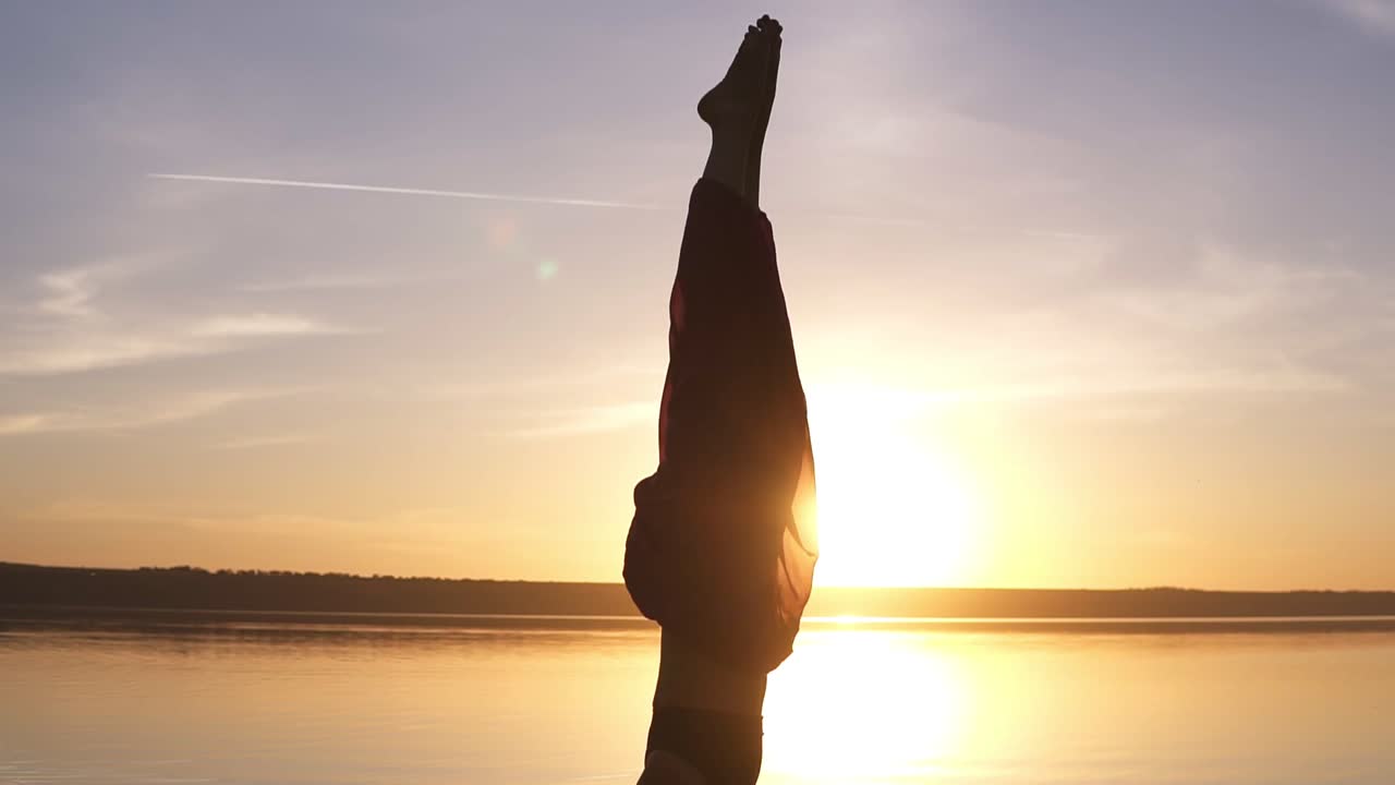 요가 아사나에서 머리에 서 있는 아름다운 소녀의 근접 영상. 태양이 빛나고 배경에 물이 빛납니다.