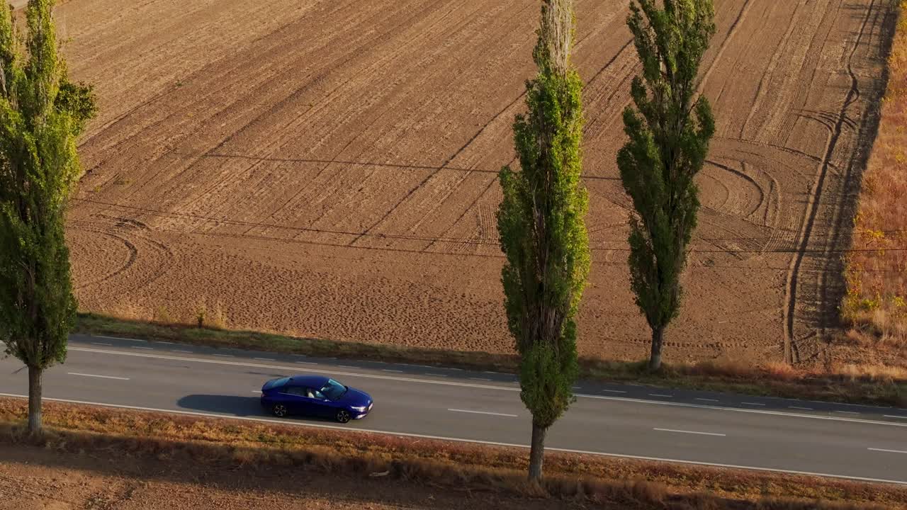 Ampia ripresa con drone in movimento di un'auto blu che guida tra campi pianeggianti, lungo una strada asfaltata illuminata dal sole, circondata da alberi verdi in una giornata di sole.