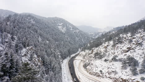 aerial reveal backwards of boulder canyon drive in colorado during the winter as cars drive down icy road surrounded by rocky mountains and snow covered pine trees