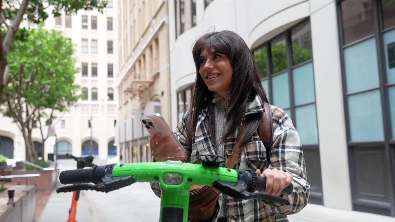 Joven sonriente usando un smartphone con un patinete eléctrico en la ciudad.