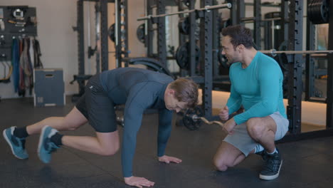 Man-Doing-Mountain-Climber-Plank-Exercise-Cheered-Up-By-Personal-Trainer-or-Gym-Friend