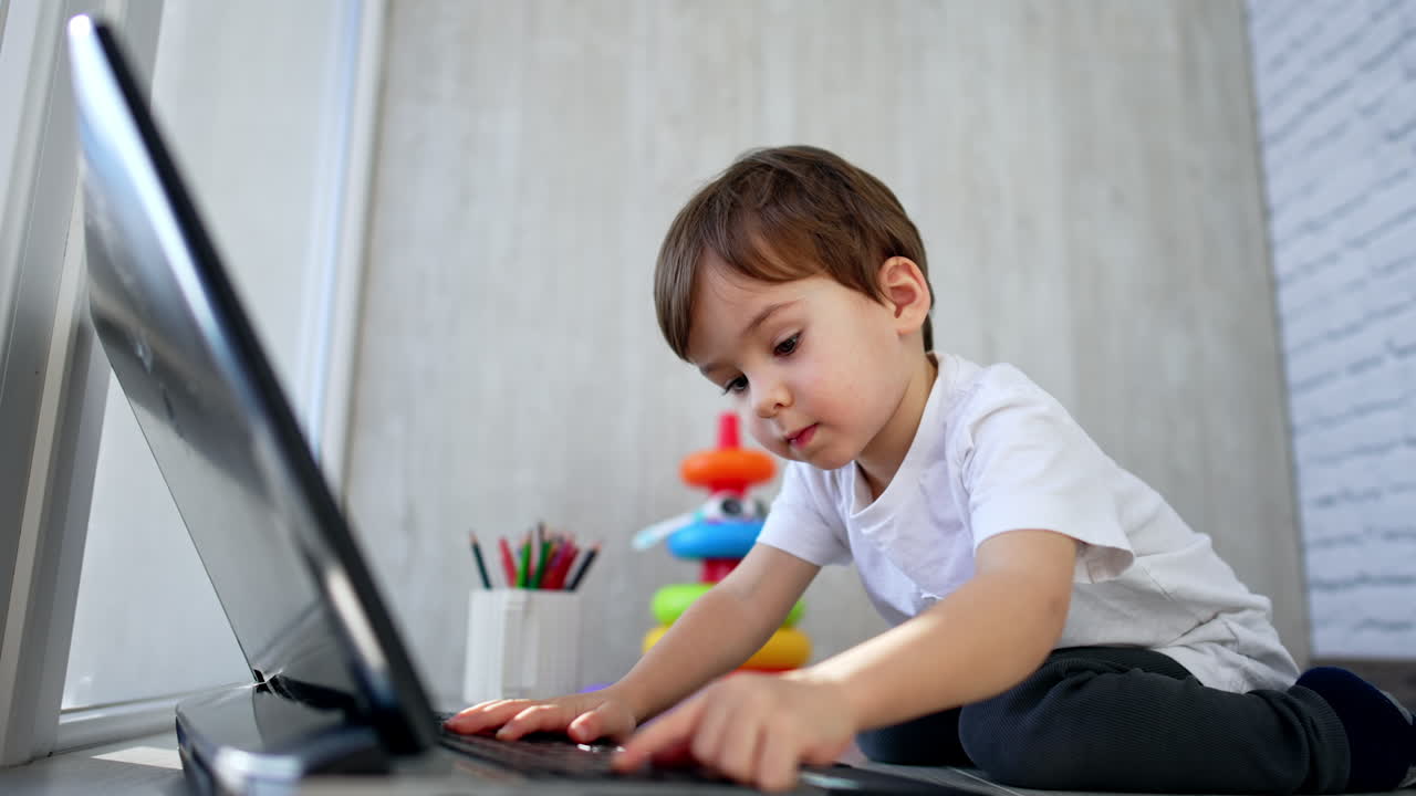 Caucasian two-year old toddler sits on the floor near laptop. Side view. Focused kid presses keys on the keyboard.