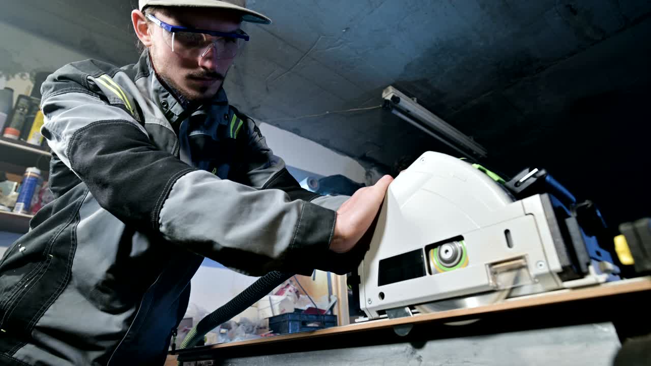 회색 오버을 입은 수염을 가진 젊은 남자가 직업으로 목수입니다. 원형 절단 기계로 일합니다.