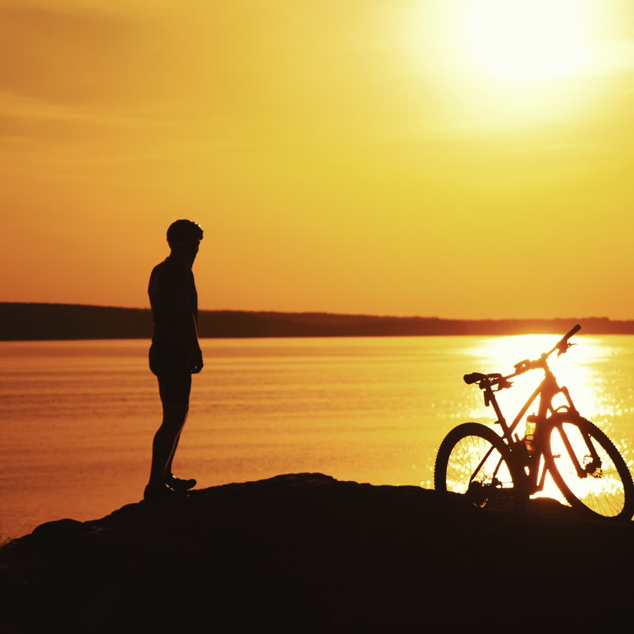 Silhouette of a sportive body of a cyclist near the river at beautiful sunset. Male biker standing next to his bike on the evening river background at amazing golden sunset.