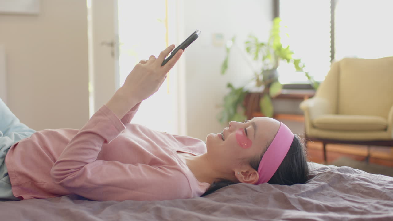 Relaxing on bed, woman with eye patches holding smartphone, resting at home