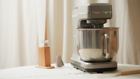electric mixer with stainless steel bowl sits on table beside wooden cake stand, piping tools, and spatula in bright kitchen setting ready for home baking preparation with natural daylight