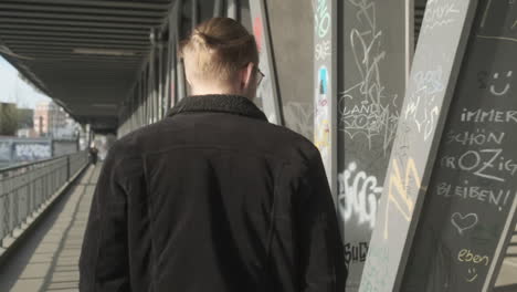 Young-European-Man-with-Man-Bun,-Glasses-and-a-Black-Jacket-Walking-on-Sidewalk-during-Daytime-in-Hamburg,-Germany