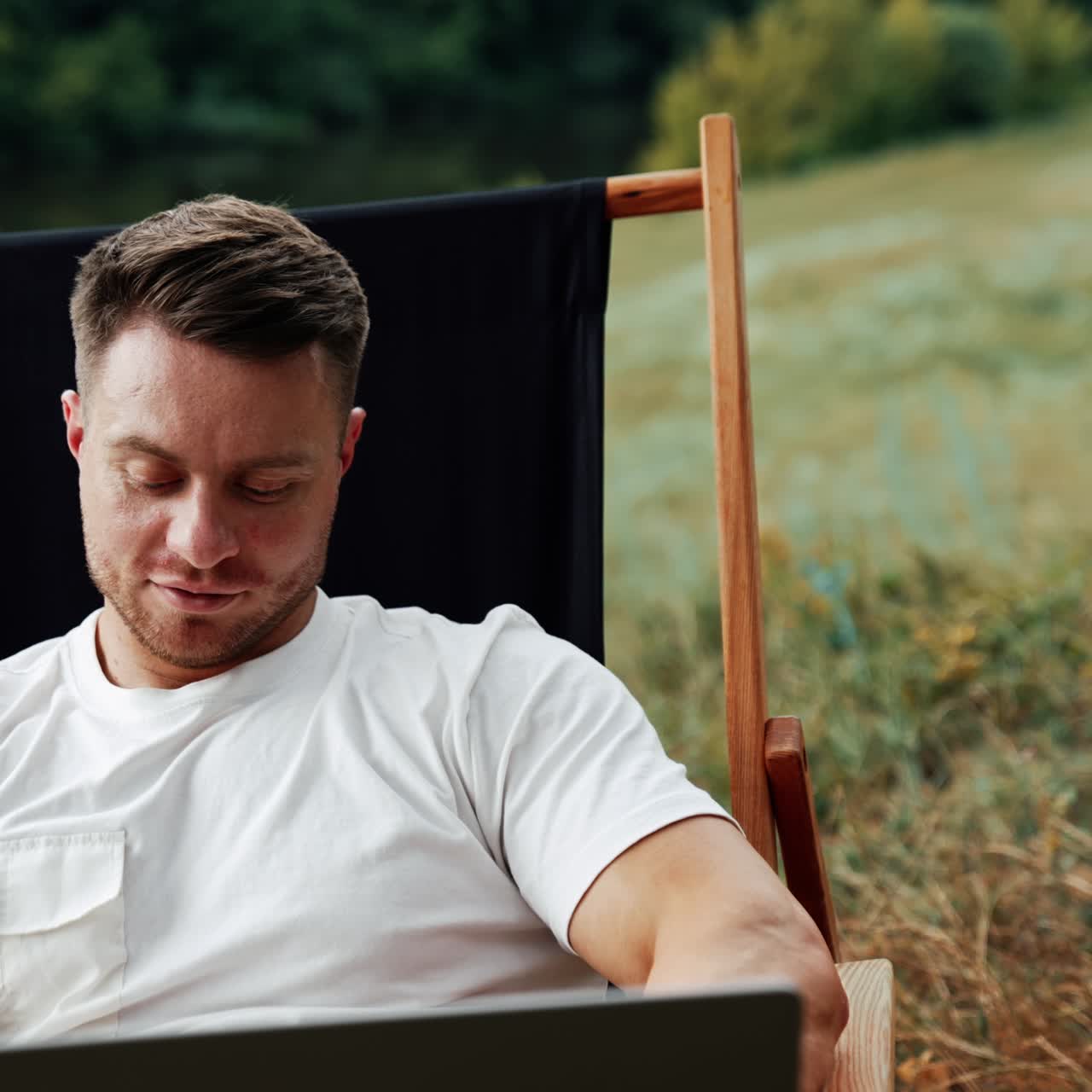 Cheerful Caucasian entrepreneur looks at his laptop. Man working relaxed in the nature backdrop