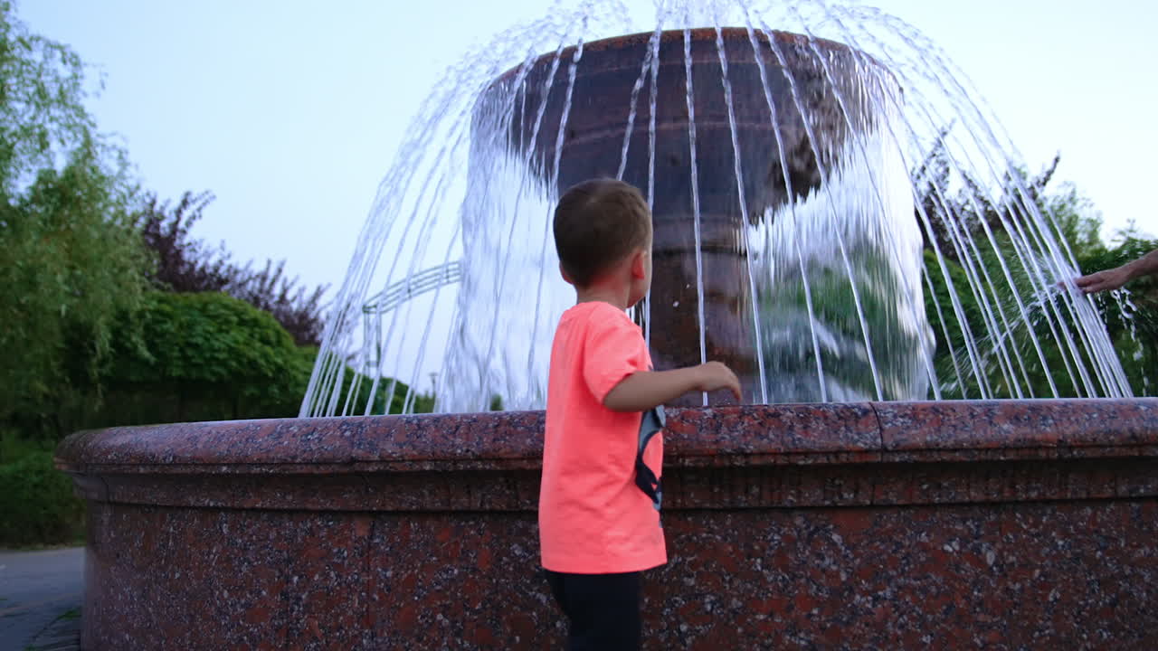 Lovely baby stands at the fountain in the park. Happy energetic kid jumps cheerfully looking at water. Low angle view.