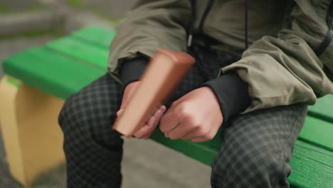 close up partial view of person in checkered pants sitting on green bench holding brown cover novel in left hand with gesture suggesting thoughtful hitting motion