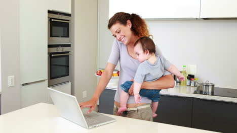 mother holding her happy baby boy and talking on phone using laptop