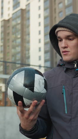 hombre de pie gira la pelota de fútbol en su palma con precisión, enfocándose atentamente, en un fondo urbano con edificios de gran altura y cercas de malla metálica