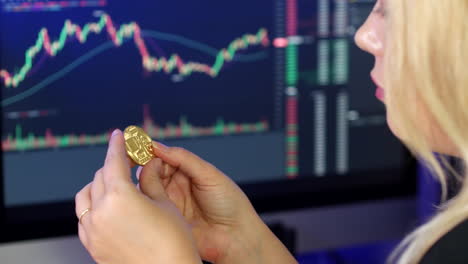 blonde woman holding and looking at bitcoin in her hands in front of cryptocurrency trading graphs