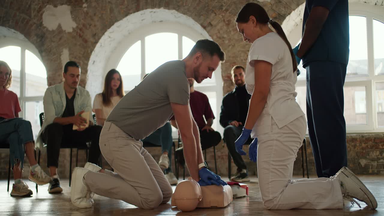 회색 티셔츠를 입은 수염이 있는 갈색 남자가 인공 호흡을 시키고, 색 유니폼을 입은 간호사 소녀가 벽돌 벽과 큰 창문을 배경으로 대중 앞에서 인공 수술을 시킨다.